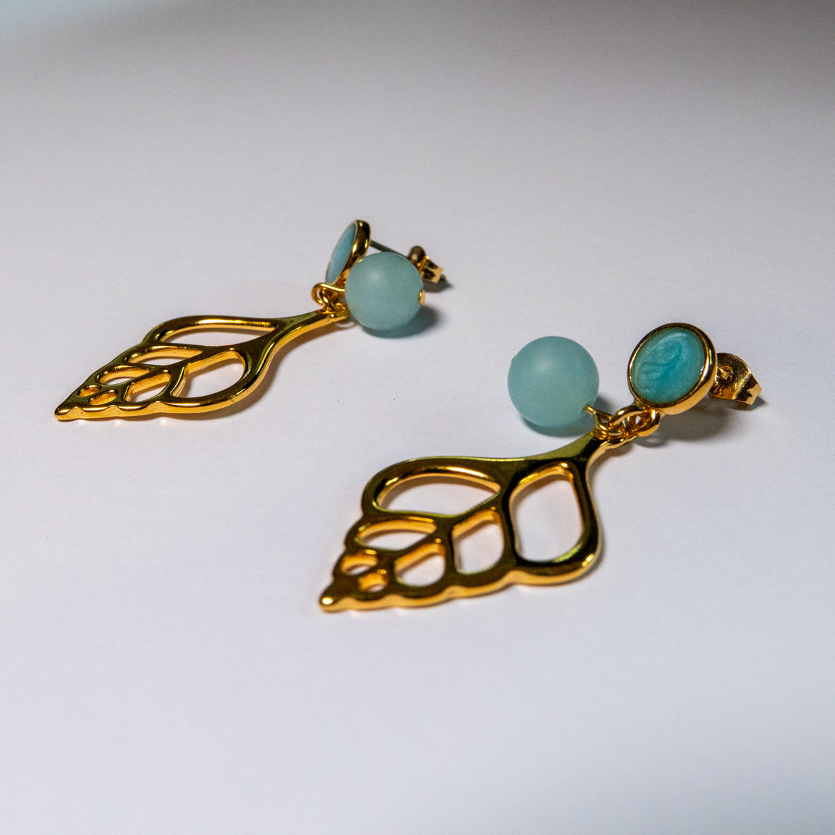 Sunlit Shell oorbellen met matte turquoise kralen en opengewerkte goudkleurige schelphangers op een witte achtergrond.