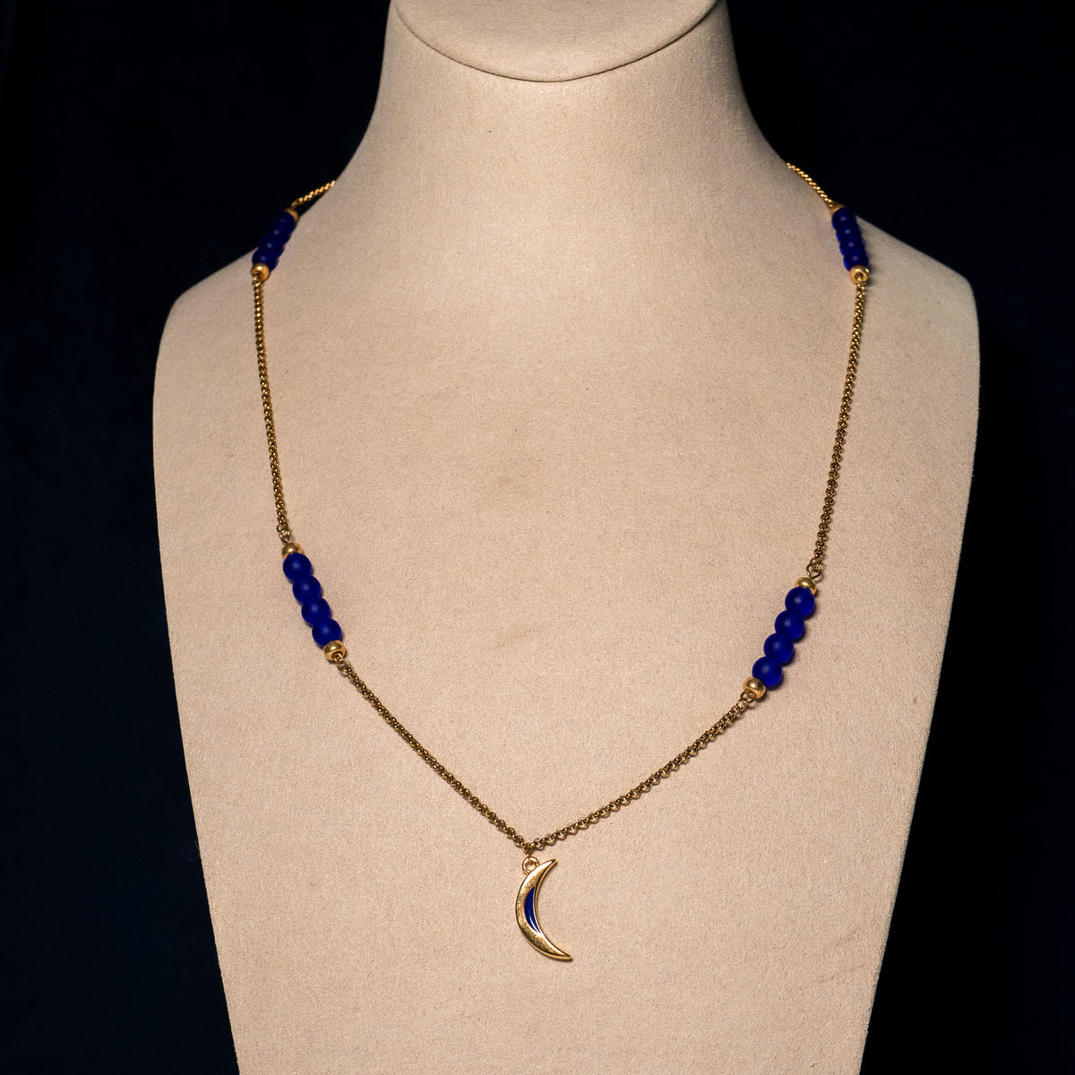 Midnight Moon ketting op beige buste. Gouden maan met blauw detail aan goudkleurige ketting met blauwe glaskralen.