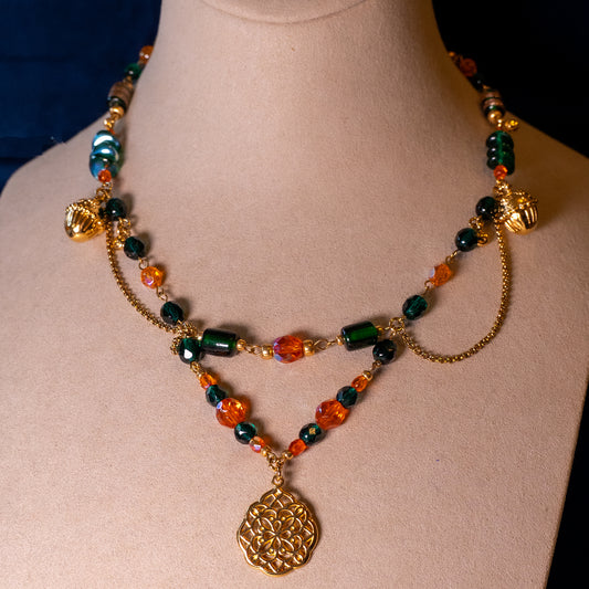 De ketting Oakheart op een beige buste. Oranje en groene glaskralen gecombineerd met goudkleurige hangende elementen en basismaterialen.