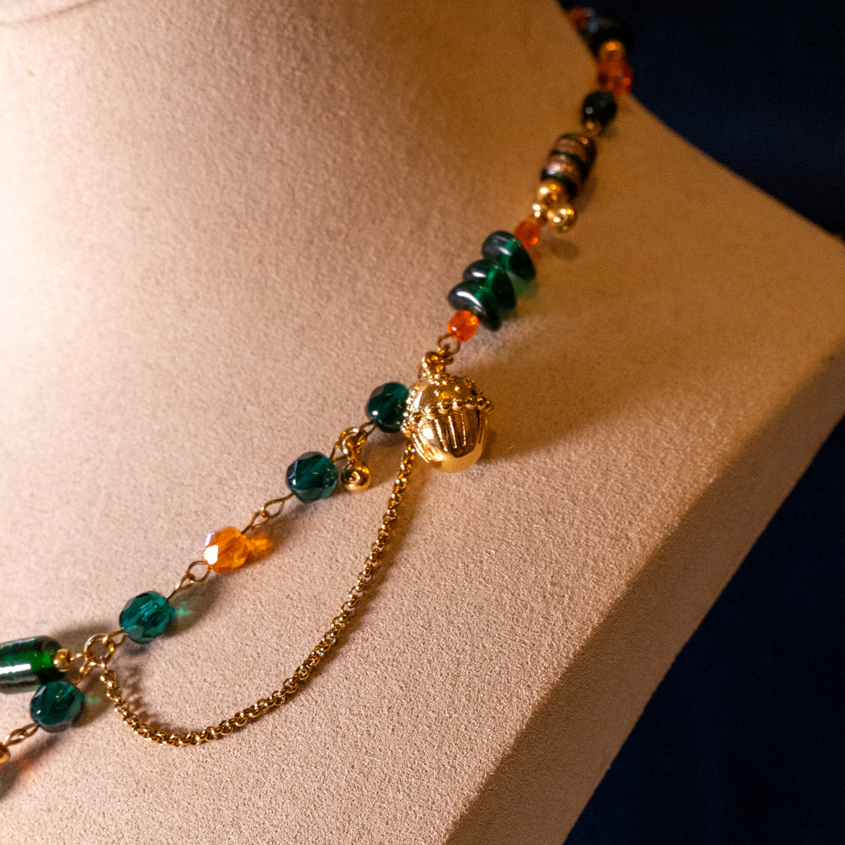 De ketting Oakheart op een beige buste. Oranje en groene glaskralen gecombineerd met goudkleurige hangende elementen en basismaterialen. Detailfoto van eikel.
