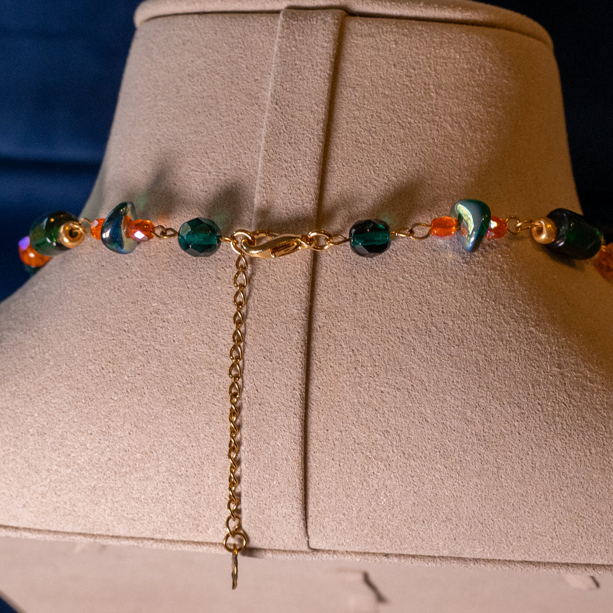 De ketting Oakheart op een beige buste. Oranje en groene glaskralen gecombineerd met goudkleurige hangende elementen en basismaterialen. Detailfoto van sluiting.