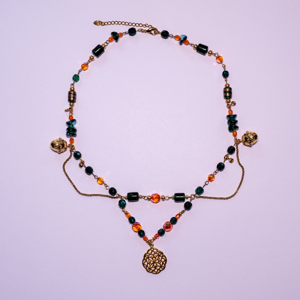 De ketting Oakheart op een witte achtergond Oranje en groene glaskralen gecombineerd met goudkleurige hangende elementen en basismaterialen.