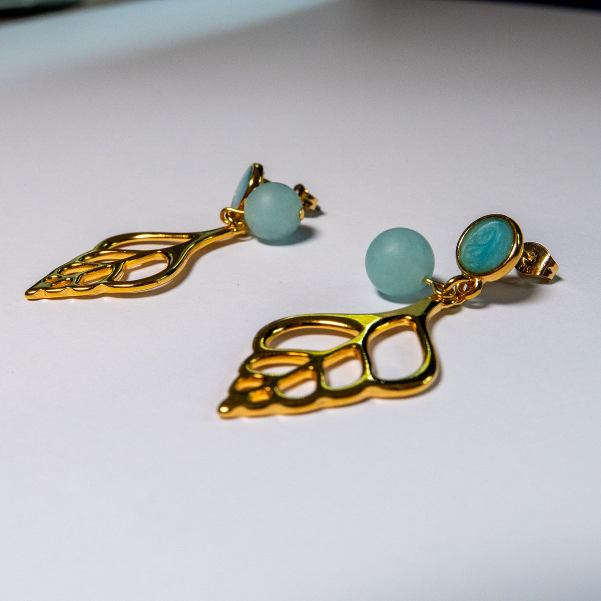 Sunlit Shell oorbellen met matte turquoise kralen en opengewerkte goudkleurige schelphangers op een witte achtergrond.
